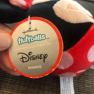 Op de afbeelding: Een rood en wit pluche Minnie Mouse-ornament met zwarte accenten. Het ornament is een Hallmark fluffball en heeft een label met de tekst "Hallmark fluffballs TM/MC Ornament/Decoration Disney Minnie".