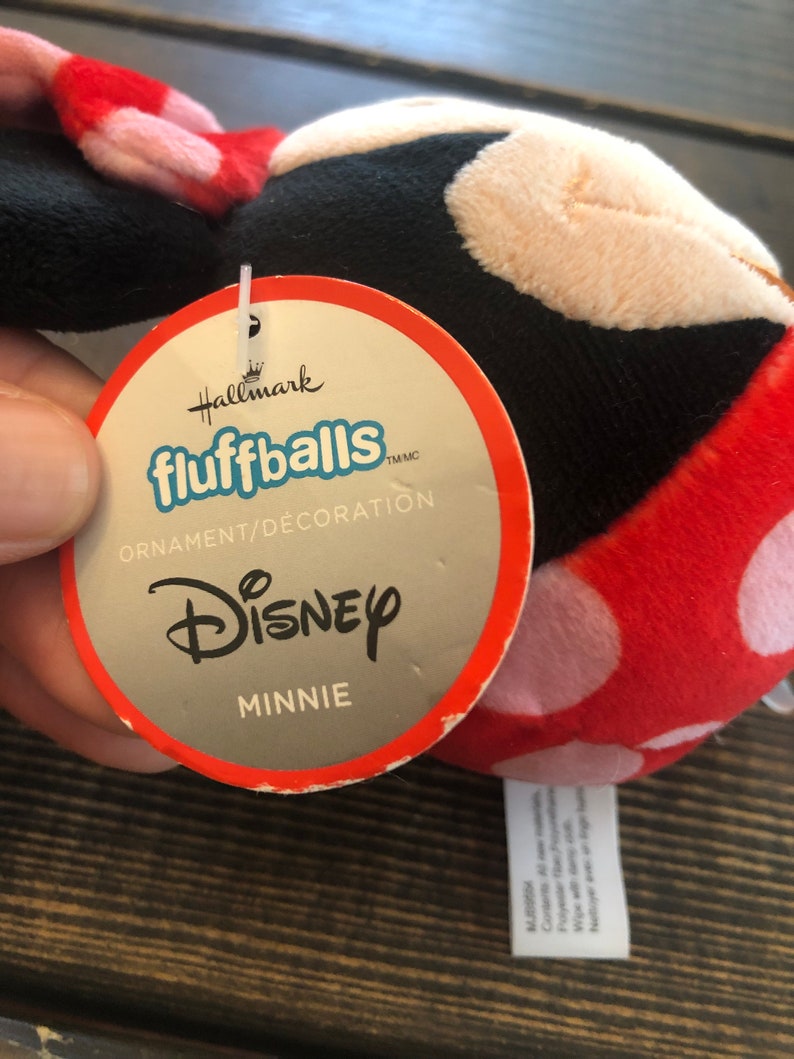 Op de afbeelding: Een rood en wit pluche Minnie Mouse-ornament met zwarte accenten. Het ornament is een Hallmark fluffball en heeft een label met de tekst "Hallmark fluffballs TM/MC Ornament/Decoration Disney Minnie".