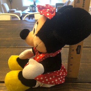May include: A plush toy of Minnie Mouse sitting with her legs crossed. She is wearing a red dress with white polka dots and a red bow with white polka dots. She has black ears and a yellow shoe on her left foot.