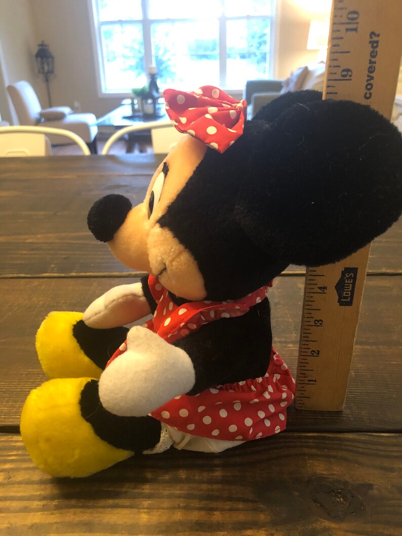 May include: A plush toy of Minnie Mouse sitting with her legs crossed. She is wearing a red dress with white polka dots and a red bow with white polka dots. She has black ears and a yellow shoe on her left foot.