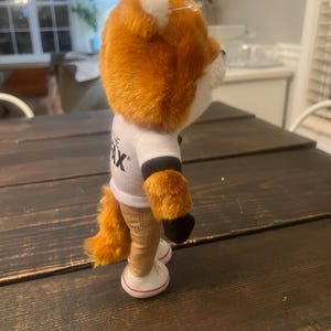 May include: A plush fox toy with orange fur, white accents, and a white t-shirt with black sleeves. The fox wears tan trousers and white trainers with red trim. The toy is standing on a dark wooden surface.