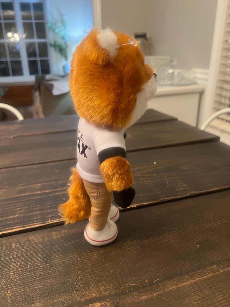 May include: A plush fox toy with orange fur, white accents, and a white t-shirt with black sleeves. The fox wears tan trousers and white trainers with red trim. The toy is standing on a dark wooden surface.