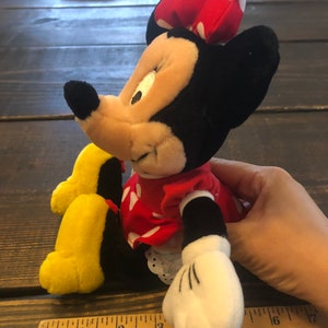 May include: A plush Minnie Mouse toy with a red dress with white polka dots, a red bow with white polka dots, and yellow shoes. The toy is sitting on a wooden surface with a ruler.