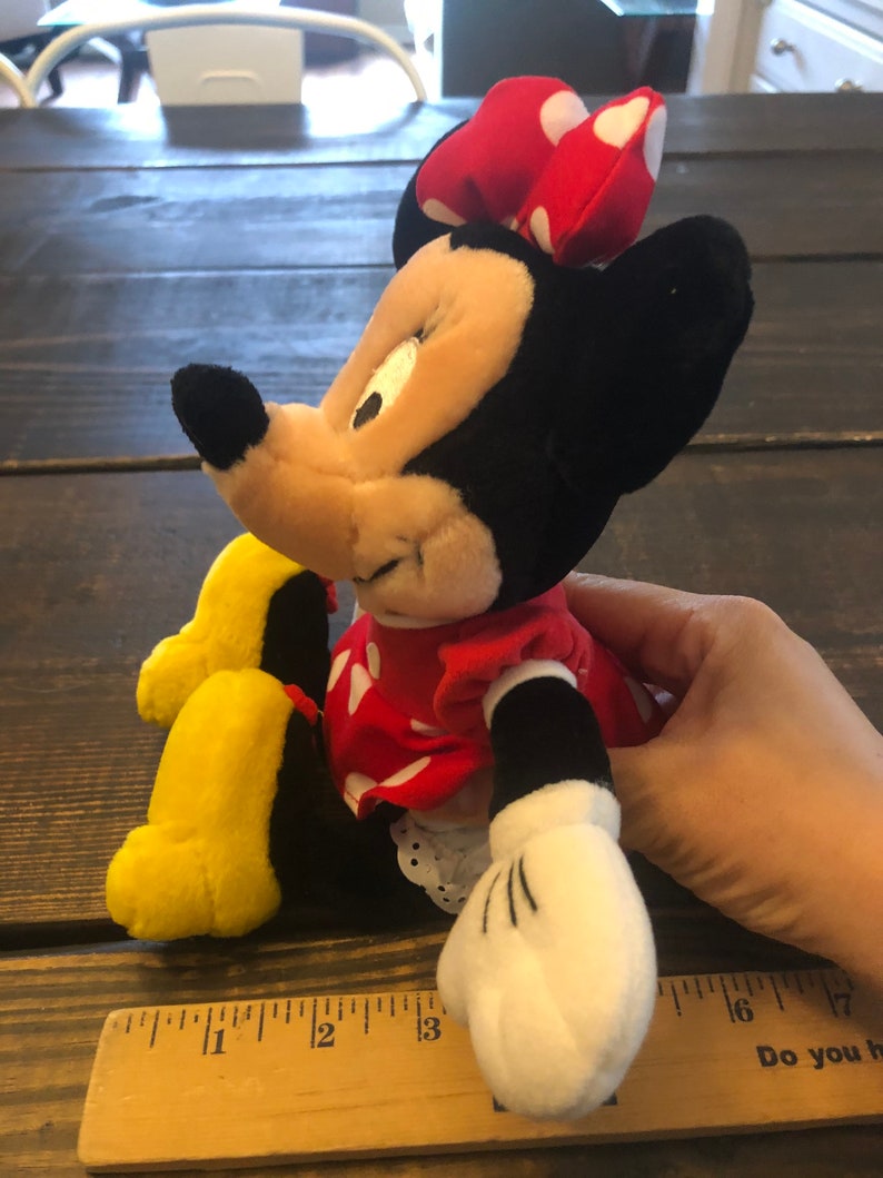May include: A plush Minnie Mouse toy with a red dress with white polka dots, a red bow with white polka dots, and yellow shoes. The toy is sitting on a wooden surface with a ruler.