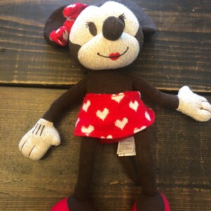 K&ouml;nnte beinhalten: Ein gestricktes Pl&uuml;schtier Minnie Mouse mit einem rot-wei&szlig; gepunkteten Rock und einer roten Schleife. Das Spielzeug hat braune Augen, eine braune Nase und rote Lippen.