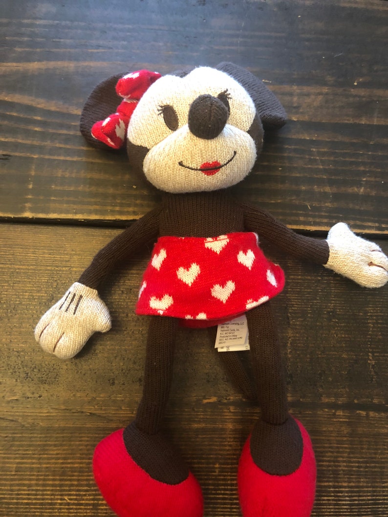 K&ouml;nnte beinhalten: Ein gestricktes Pl&uuml;schtier Minnie Mouse mit einem rot-wei&szlig; gepunkteten Rock und einer roten Schleife. Das Spielzeug hat braune Augen, eine braune Nase und rote Lippen.