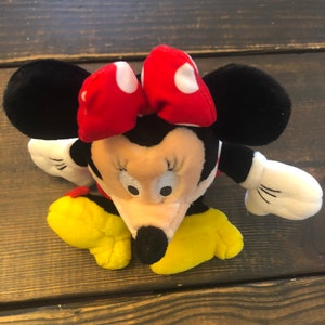 May include: A plush toy of Minnie Mouse, with black ears, a red bow with white polka dots, a black nose, and yellow shoes.