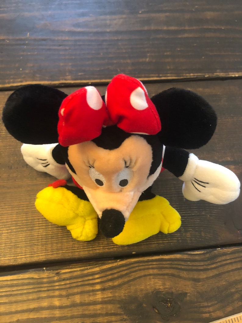 May include: A plush toy of Minnie Mouse, with black ears, a red bow with white polka dots, a black nose, and yellow shoes.