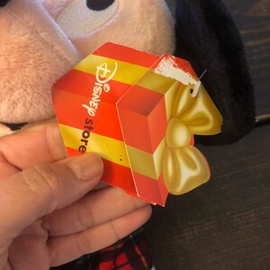 May include: A plush pink and black Mickey Mouse toy wearing a red and black plaid outfit. The toy has a gift tag attached that says "Disney store".