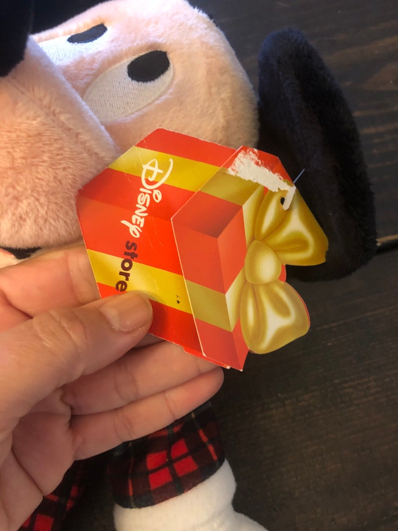 May include: A plush pink and black Mickey Mouse toy wearing a red and black plaid outfit. The toy has a gift tag attached that says "Disney store".