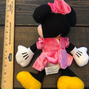 May include: A pink and black plush Minnie Mouse toy wearing a pink dress and a pink bow. The toy has yellow shoes and black eyes. The toy is made in China.