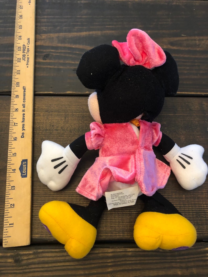 May include: A pink and black plush Minnie Mouse toy wearing a pink dress and a pink bow. The toy has yellow shoes and black eyes. The toy is made in China.
