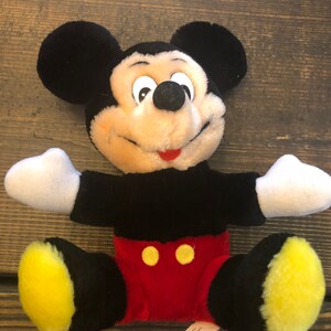 May include: A black and yellow plush Mickey Mouse toy sitting with its arms outstretched. The toy has a red and yellow outfit and a tag that reads "All new material, Polyester stuffing, MFR. 105, Made in Korea."
