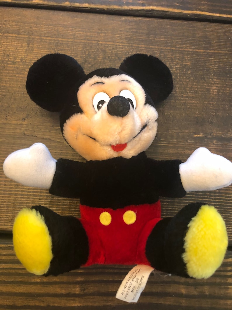 May include: A black and yellow plush Mickey Mouse toy sitting with its arms outstretched. The toy has a red and yellow outfit and a tag that reads "All new material, Polyester stuffing, MFR. 105, Made in Korea."