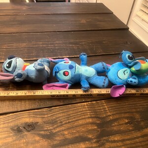 Op de afbeelding: Drie blauwe Stitch-knuffels met grote oren en opvallende kenmerken zijn gerangschikt op een houten oppervlak. E&eacute;n speelgoed heeft een kleine, groene en blauwe schildpad op zijn rug. Een houten liniaal staat op de voorgrond.
