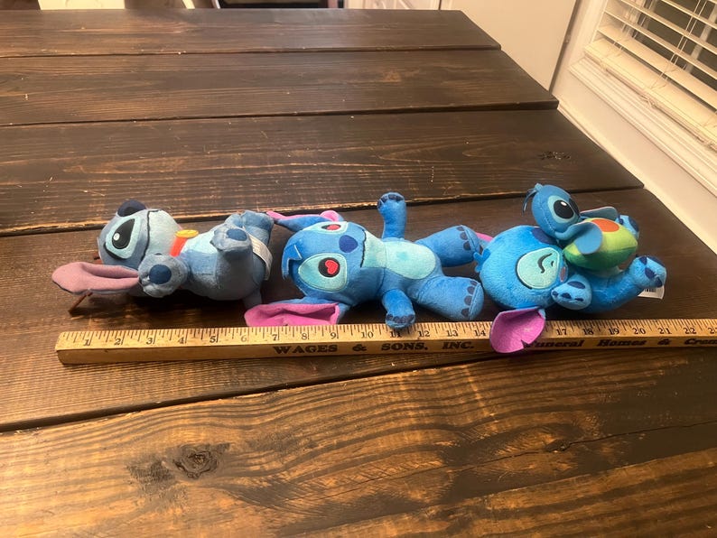 Op de afbeelding: Drie blauwe Stitch-knuffels met grote oren en opvallende kenmerken zijn gerangschikt op een houten oppervlak. E&eacute;n speelgoed heeft een kleine, groene en blauwe schildpad op zijn rug. Een houten liniaal staat op de voorgrond.