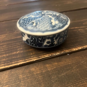 May include: A round, blue and white ceramic trinket box with a lid. The lid features a detailed pattern of floral and geometric designs. The box is set on a dark wooden surface, showcasing its intricate details and craftsmanship.