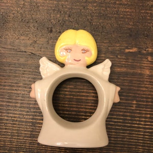 Puede incluir: Un anillo de servilleta de cer&aacute;mica con forma de &aacute;ngel con cabello rubio, vestido blanco y alas. El &aacute;ngel tiene una cara sonriente con ojos azules.