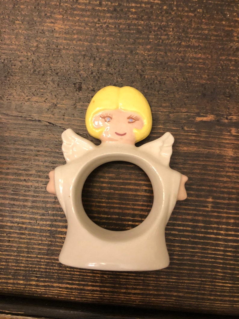 Puede incluir: Un anillo de servilleta de cer&aacute;mica con forma de &aacute;ngel con cabello rubio, vestido blanco y alas. El &aacute;ngel tiene una cara sonriente con ojos azules.