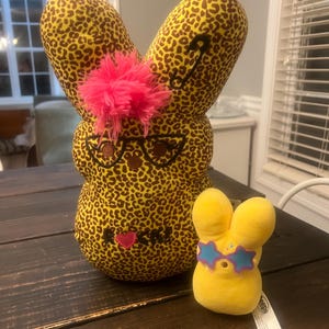 May include: Two plush bunny figures. One is large, with a leopard print pattern, pink hair, and glasses. The other is small, yellow, and wearing star-shaped sunglasses. Both are on a dark wooden surface.