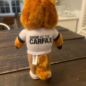 May include: A plush fox toy with orange fur, wearing a white t-shirt with the text "SHOW ME THE CARFAX". The fox has brown trousers, a fluffy tail, and white-soled shoes. The toy is on a dark wooden surface.