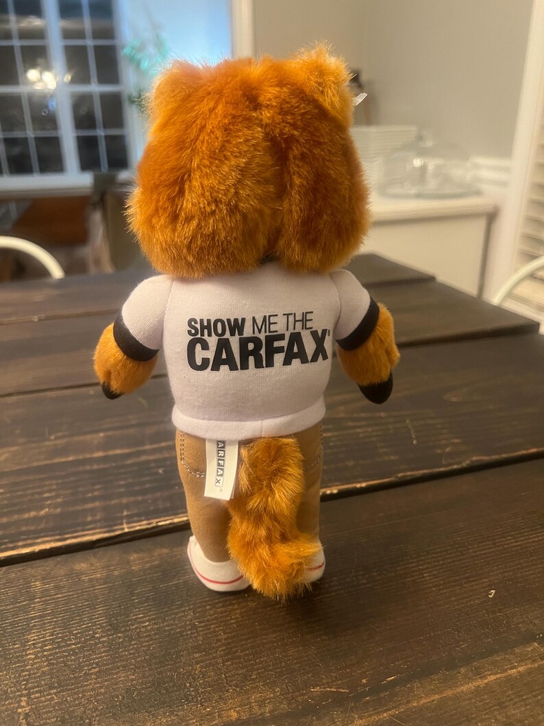 May include: A plush fox toy with orange fur, wearing a white t-shirt with the text "SHOW ME THE CARFAX". The fox has brown trousers, a fluffy tail, and white-soled shoes. The toy is on a dark wooden surface.