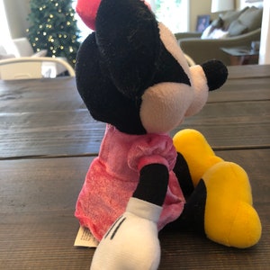 May include: A plush toy of Minnie Mouse wearing a pink dress and yellow shoes. She is sitting on a wooden surface.
