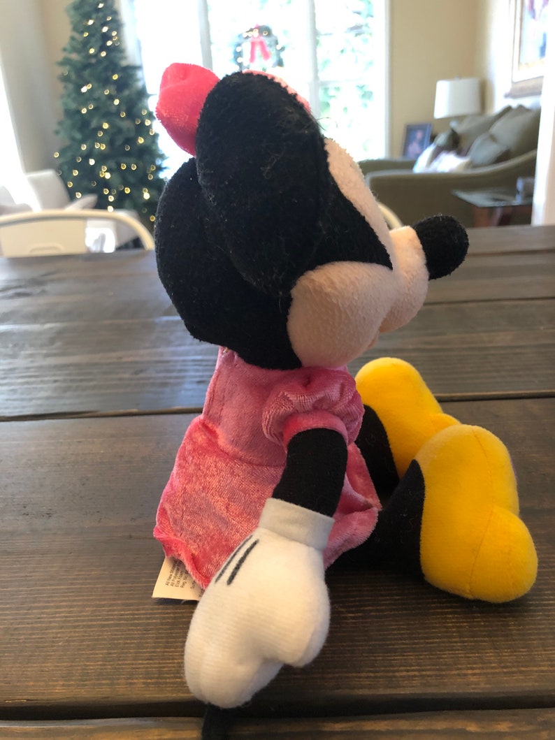 May include: A plush toy of Minnie Mouse wearing a pink dress and yellow shoes. She is sitting on a wooden surface.