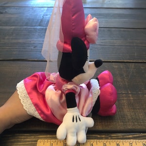 May include: Pink plush Minnie Mouse wearing a pink dress and a white veil. She is sitting with her right hand visible. The plush toy is about 12 inches tall.