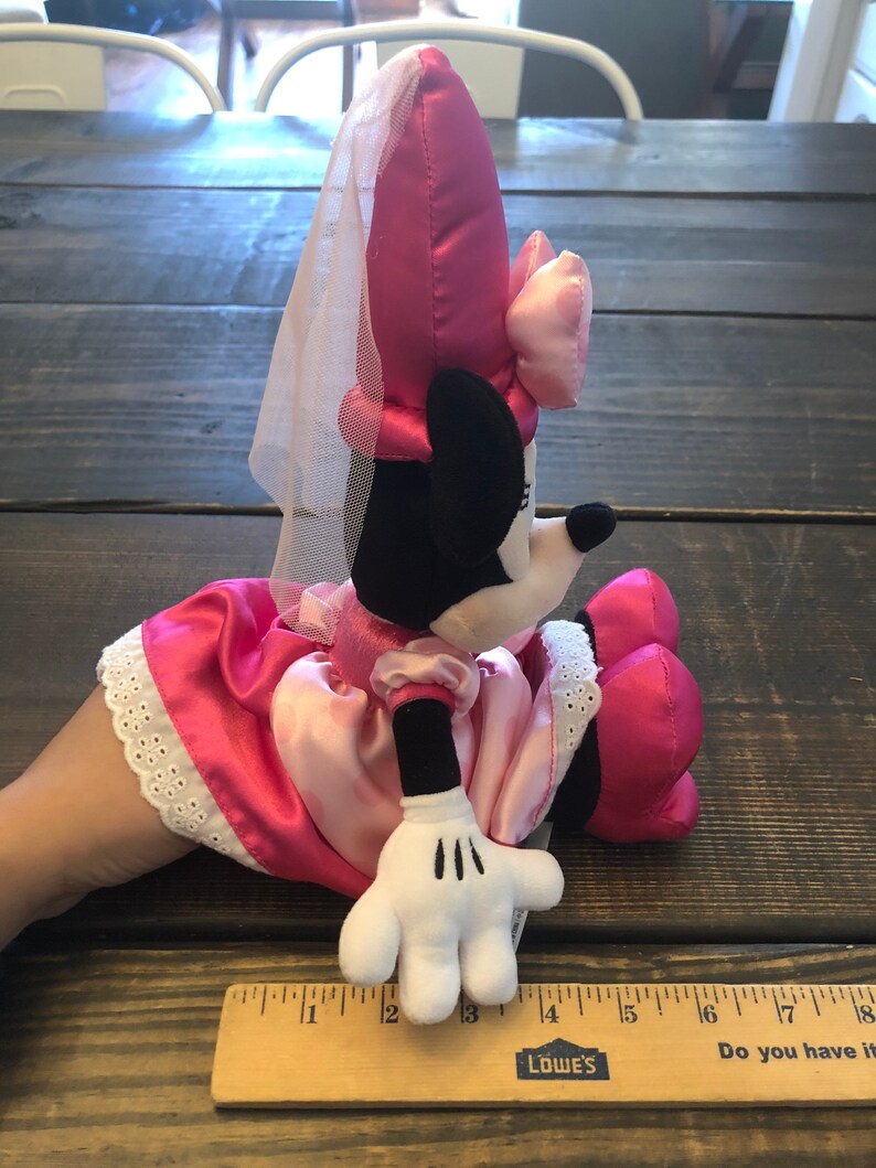 May include: Pink plush Minnie Mouse wearing a pink dress and a white veil. She is sitting with her right hand visible. The plush toy is about 12 inches tall.