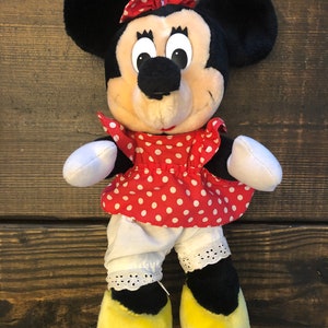 K&ouml;nnte beinhalten: Ein Pl&uuml;schtier von Minnie Mouse, das ein rotes Kleid mit wei&szlig;en Punkten, einen wei&szlig;en Petticoat mit Spitzenbesatz und gelbe Schuhe tr&auml;gt. Sie hat eine rote Schleife mit wei&szlig;en Punkten im Haar.