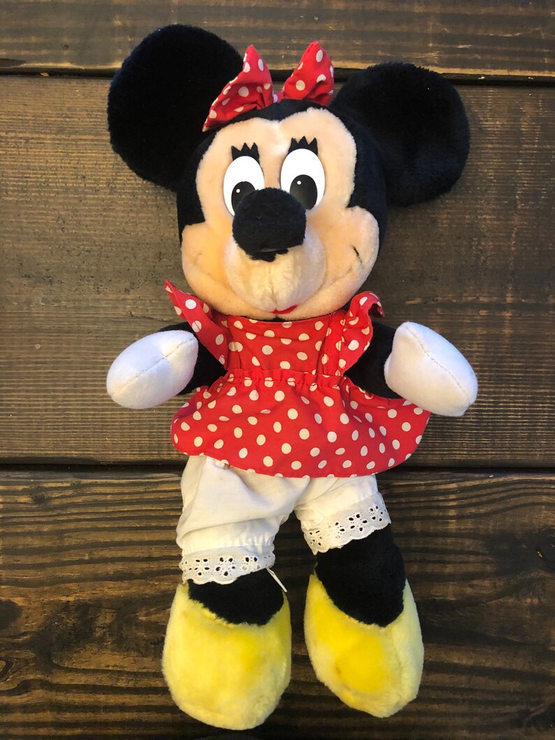 K&ouml;nnte beinhalten: Ein Pl&uuml;schtier von Minnie Mouse, das ein rotes Kleid mit wei&szlig;en Punkten, einen wei&szlig;en Petticoat mit Spitzenbesatz und gelbe Schuhe tr&auml;gt. Sie hat eine rote Schleife mit wei&szlig;en Punkten im Haar.
