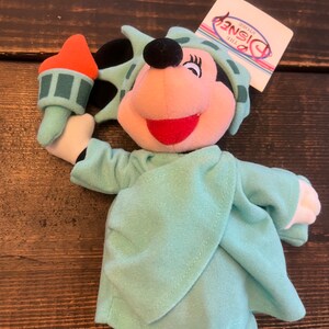 May include: A plush toy of Minnie Mouse dressed as the Statue of Liberty. She is holding a torch in her right hand and is wearing a green robe with a crown. The toy has a tag that says "The Disney Store".