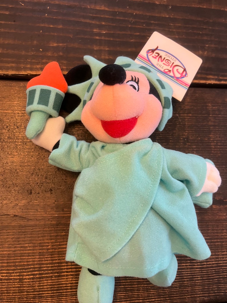 May include: A plush toy of Minnie Mouse dressed as the Statue of Liberty. She is holding a torch in her right hand and is wearing a green robe with a crown. The toy has a tag that says "The Disney Store".