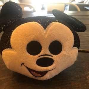 May include: A black and white plush toy of Mickey Mouse with a sparkly black fabric. The toy has a big smile and large black eyes.