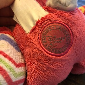 Op de afbeelding: Roze pluche speelgoed met een witte staart en een gestreept patroon op &eacute;&eacute;n been. Het speelgoed heeft een bruin geborduurd patch met de tekst "Genuine Original Disney Store" erop.