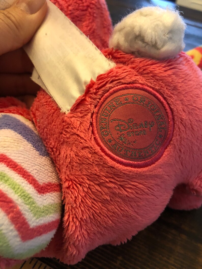 Op de afbeelding: Roze pluche speelgoed met een witte staart en een gestreept patroon op &eacute;&eacute;n been. Het speelgoed heeft een bruin geborduurd patch met de tekst "Genuine Original Disney Store" erop.