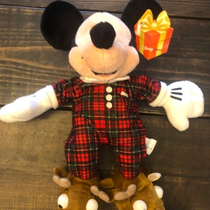 May include: A plush Mickey Mouse wearing red and green plaid pajamas and reindeer slippers. The plush toy has a Disney Store tag attached to its arm.