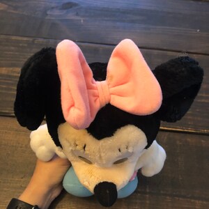 May include: A plush toy of Minnie Mouse wearing a pink bow. The toy is sitting on a wooden surface.