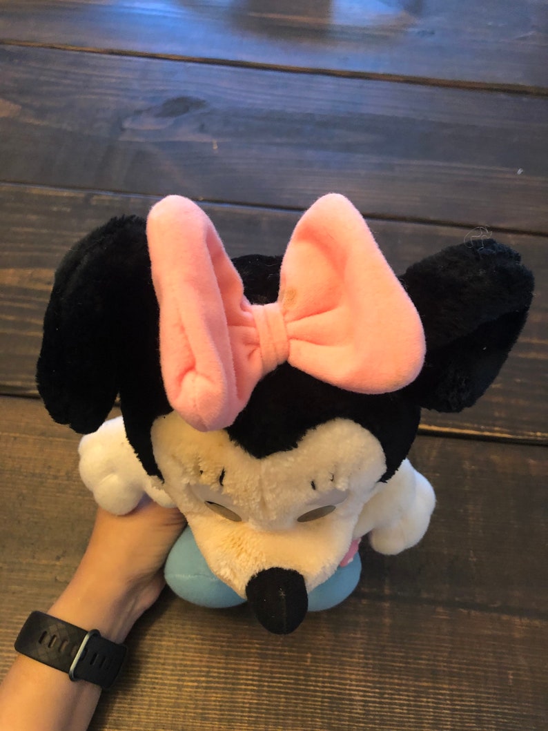 May include: A plush toy of Minnie Mouse wearing a pink bow. The toy is sitting on a wooden surface.