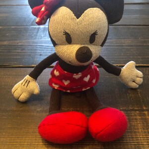 K&ouml;nnte beinhalten: Ein braunes und graues gestricktes Pl&uuml;schtier Minnie Mouse mit roten Schuhen und einer roten Schleife mit wei&szlig;en Punkten. Das Spielzeug tr&auml;gt einen roten Rock mit wei&szlig;en Herzen.