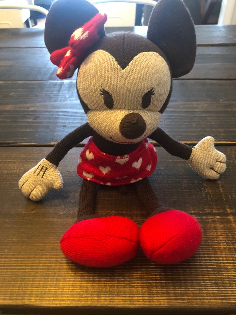 K&ouml;nnte beinhalten: Ein braunes und graues gestricktes Pl&uuml;schtier Minnie Mouse mit roten Schuhen und einer roten Schleife mit wei&szlig;en Punkten. Das Spielzeug tr&auml;gt einen roten Rock mit wei&szlig;en Herzen.