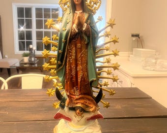 Puede incluir: Estatua detallada de una figura religiosa, que representa a una mujer con una túnica fluida con una capa verde y un halo dorado de estrellas. La estatua está sobre una base decorativa con detalles dorados, sobre una mesa de madera oscura.