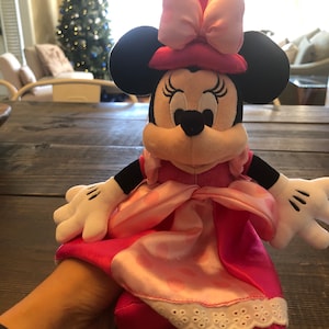 May include: Pink Minnie Mouse plush toy wearing a pink dress and a pink hat with a bow. The toy has black ears and a black nose.