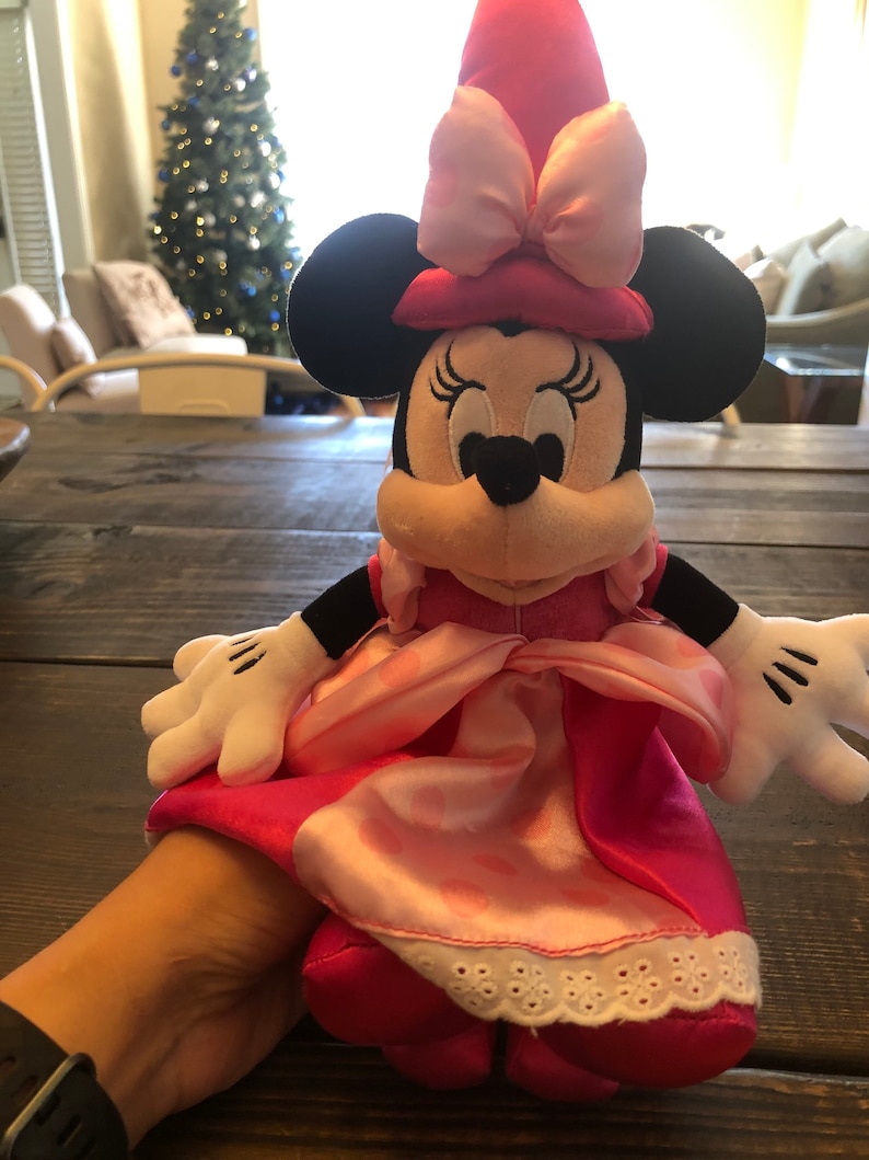 May include: Pink Minnie Mouse plush toy wearing a pink dress and a pink hat with a bow. The toy has black ears and a black nose.