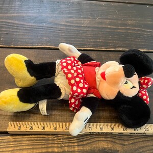 May include: A plush Minnie Mouse toy with black fur, red and white polka dot dress, and yellow shoes. The toy is lying on a wooden surface with a ruler next to it.