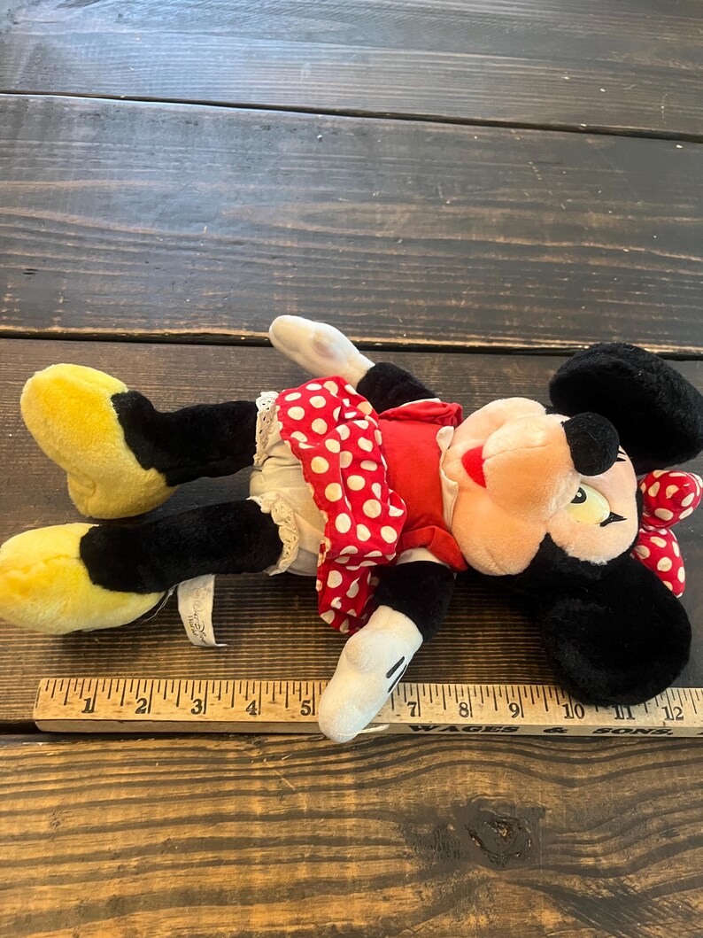 May include: A plush Minnie Mouse toy with black fur, red and white polka dot dress, and yellow shoes. The toy is lying on a wooden surface with a ruler next to it.