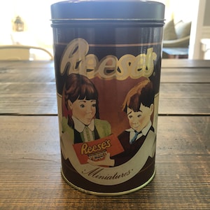 May include: A cylindrical tin of Reese's Peanut Butter Cups Miniatures. The tin is dark brown with gold lettering and illustrations of children. The Reese's logo is prominently displayed. The tin is sitting on a wooden surface.
