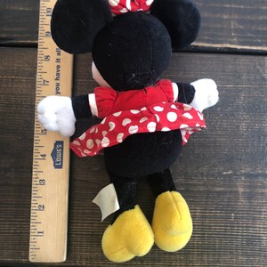 Puede incluir: Un juguete de peluche negro y rojo de Minnie Mouse que lleva un vestido rojo con lunares blancos y un lazo rojo con lunares blancos. El juguete mide aproximadamente 17 cm de alto y tiene zapatos amarillos.