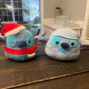 May include: Two plush toys shaped like a character with large ears. One is blue with a red Santa hat and scarf. The other is light blue with a winter hat and scarf. Both have large eyes and a dark blue nose.